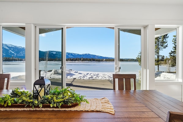 A room with a wooden table, chairs, a lantern, and a plant. It has large glass doors leading to a beach and mountains.