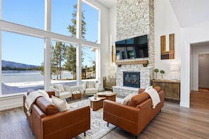 A spacious living room with a stone fireplace, large windows overlooking a lake, and modern furniture.