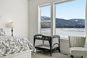 Quarto com cama, uma cadeira, janela e vista para montanhas e um lago.