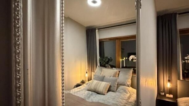 A bedroom with a large mirror, a bed with pillows, a nightstand, and a window with curtains. - Villa Bäckagård Bed & Breakfast (Åstorp)