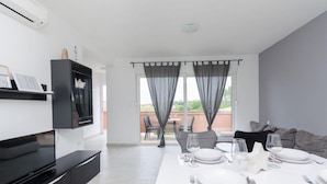 A modern dining area with a white table set for four, a flat-screen TV mounted on the wall, and a sliding glass door leading to an outdoor patio. - Apartments and Room Ljubica i Branko (Lopar)