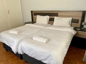 Una cama doble con ropa de cama blanca y dos toallas, un cabecero de madera, una mesita de noche con lámpara y suelo de madera.