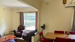 A room with a sofa, a dining table with red chairs, a fan, and a window with curtains.