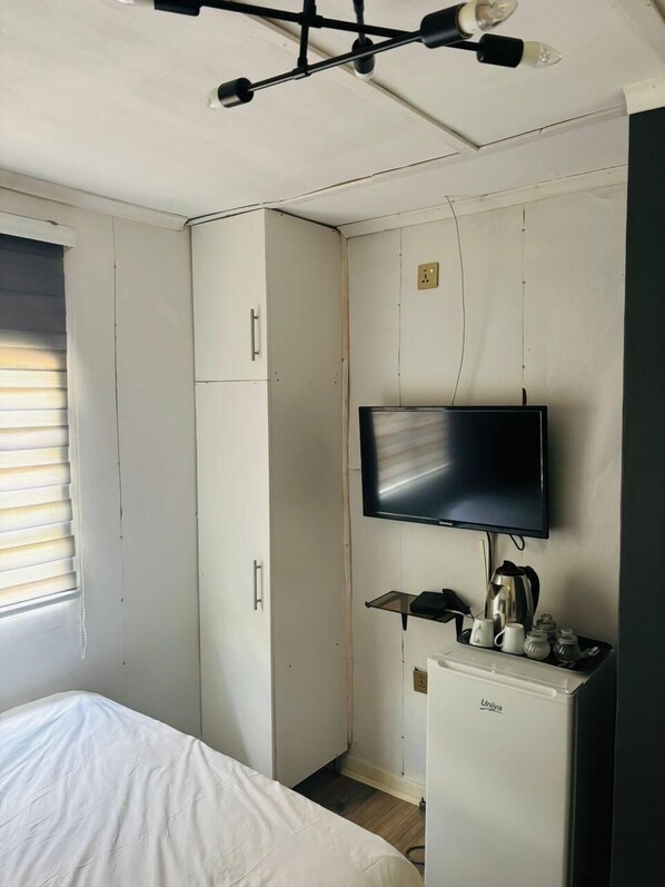 A compact hotel room with a bed, a television mounted on the wall, a small kitchenette with a coffee maker, and a wardrobe.