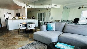 A modern living room with a grey sofa, a dining area with a round table and chairs, a kitchen with white cabinets, and a large window. - Modern & Spacious 1 br + bonus rm 2 bthrm apartment in Marina District high rise (San Diego)