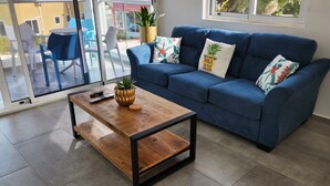 A living room with a blue sofa, a wooden coffee table, and a sliding glass door leading to an outdoor area with chairs and a plant.