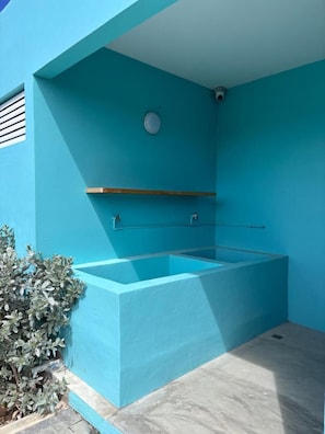 A blue-tiled outdoor pool with a wooden shelf, a metal railing, and a security camera on the wall.