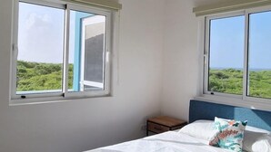 A bedroom with a bed, two windows, a nightstand, and a view of greenery.