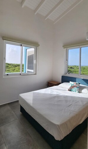 Une chambre à coucher avec un lit, deux fenêtres, une table de chevet et une vue sur la verdure.