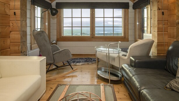 A cozy cabin interior with a large window overlooking the ocean, a leather sofa, a rocking chair, and a small table with a metal tray. - The Wooden House, Breanish Uig, Isle of Lewis (Uig)