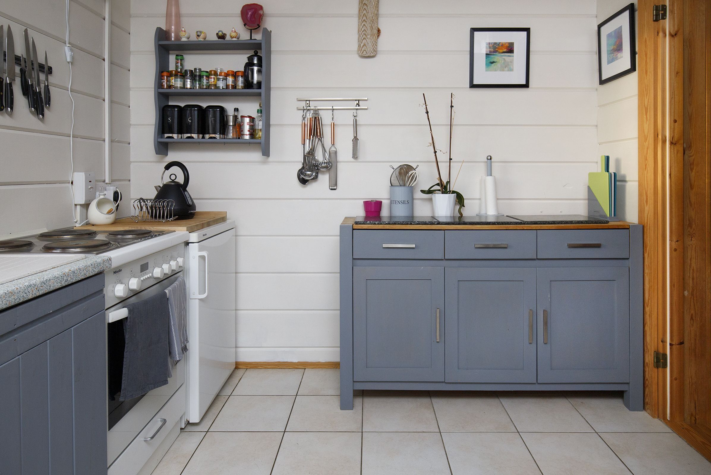 A kitchen with a stove, oven, and cabinets. There are utensils hanging on the wall and a shelf with jars and a kettle.