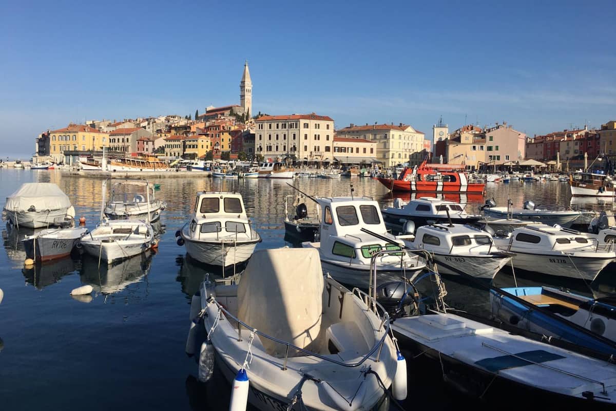 Un port de plaisance où sont amarrés plusieurs petits bateaux, une imposante tour de l’horloge en arrière-plan et une rangée de bâtiments colorés le long du quai.