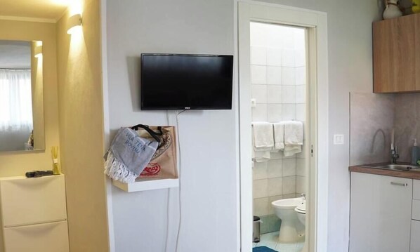 Een kleine hotelkamer met een aan de muur bevestigde televisie, een badkamer met toilet en wastafel, en een houten kastje boven de wastafel.