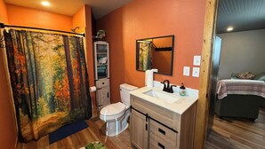 Une salle de bain avec un rideau de douche représentant une scène de forêt, des toilettes, un lavabo avec un miroir et un lit dans la pièce adjacente.