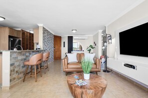 A modern kitchen with a built-in refrigerator, wooden cabinets, and a stone backsplash. There is a dining area with a round wooden table and two bar stools. A living room with a brown leather sofa and a large flat-screen TV is visible in the background. - Peña Mar Club by Otium (Fajardo)