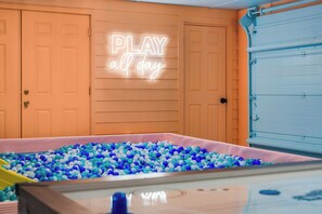 A room with a neon sign that reads "PLAY all day" above a ball pit filled with blue and white balls.
