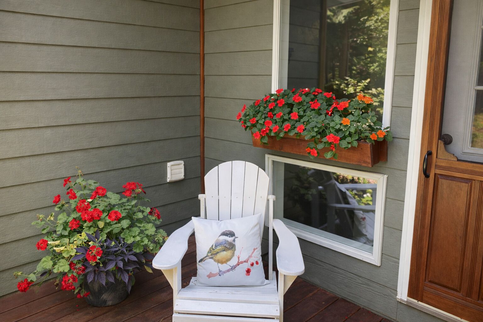 A white chair with a bird pillow on a porch with two flower planters, one with red flowers and one with orange flowers.