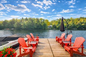 A wooden deck with red Adirondack chairs and a boat docked at the edge of a lake, surrounded by trees and a clear sky. - Moon River Cottage in Bala | BBQ, Canoes & Kayaks (MacTier)