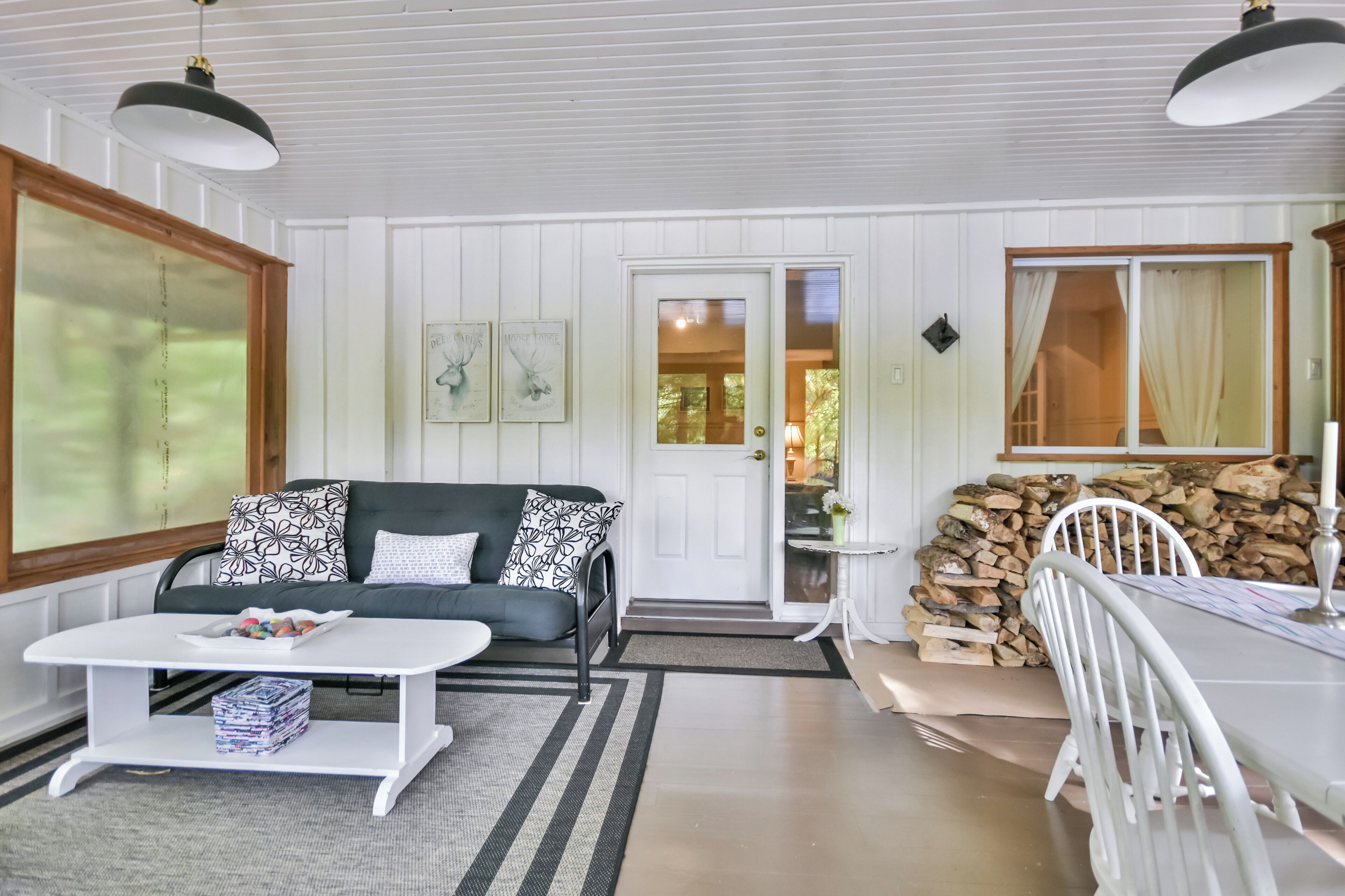 A cozy living room with a white sofa, a coffee table, and a dining table with chairs. There is a door leading to another room, a window with curtains, and a stack of firewood.