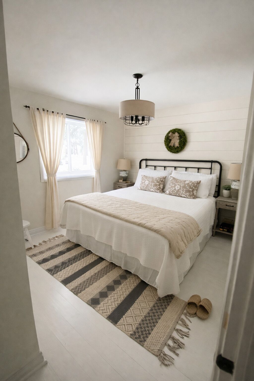 A bedroom with a bed, nightstands, a lamp, a hanging light fixture, a window with curtains, a rug, and a pair of shoes.