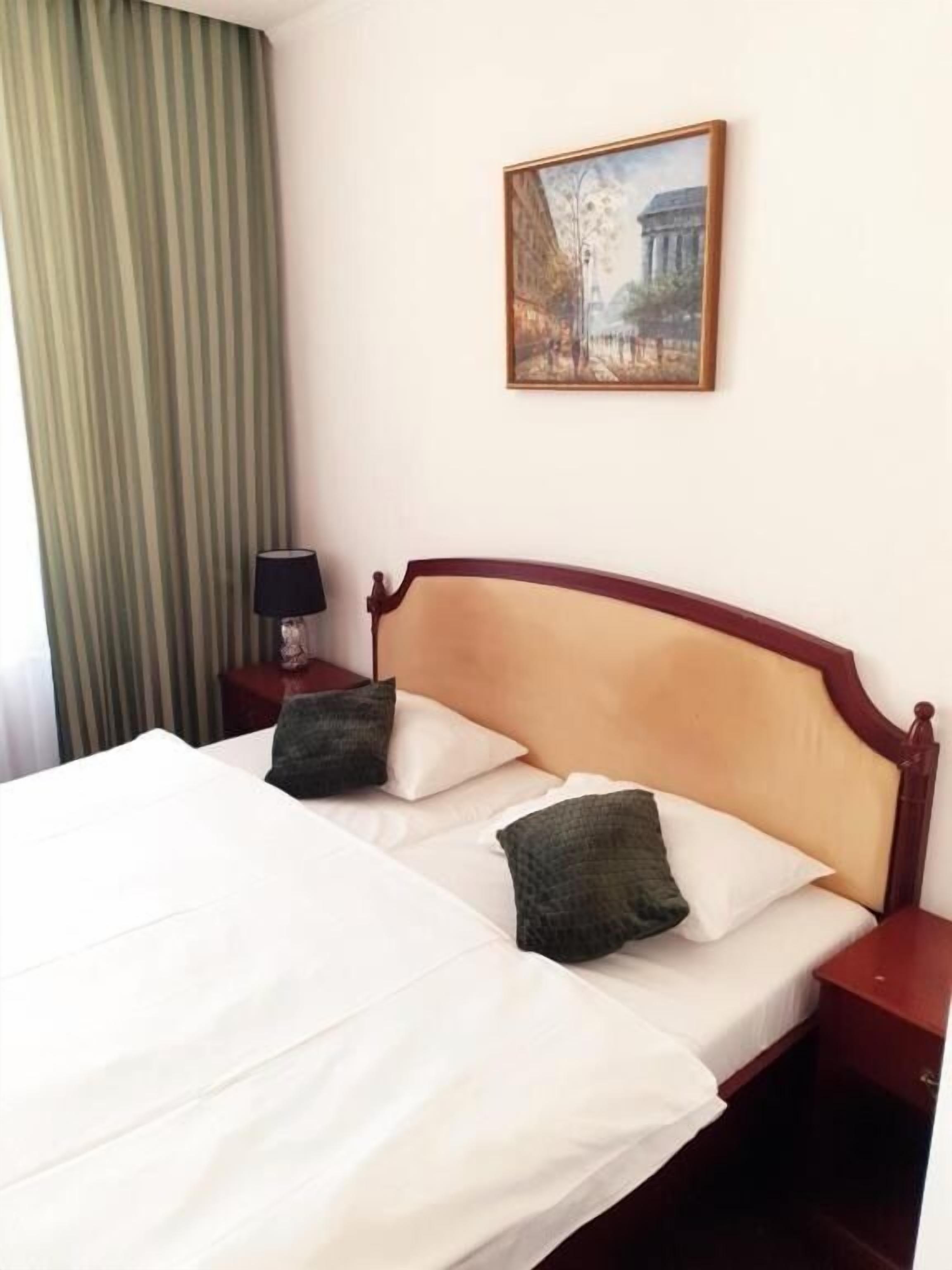 Una cama con sábanas blancas y dos almohadas, una mesita de noche con una lámpara, una cortina verde a rayas y un cuadro en la pared.