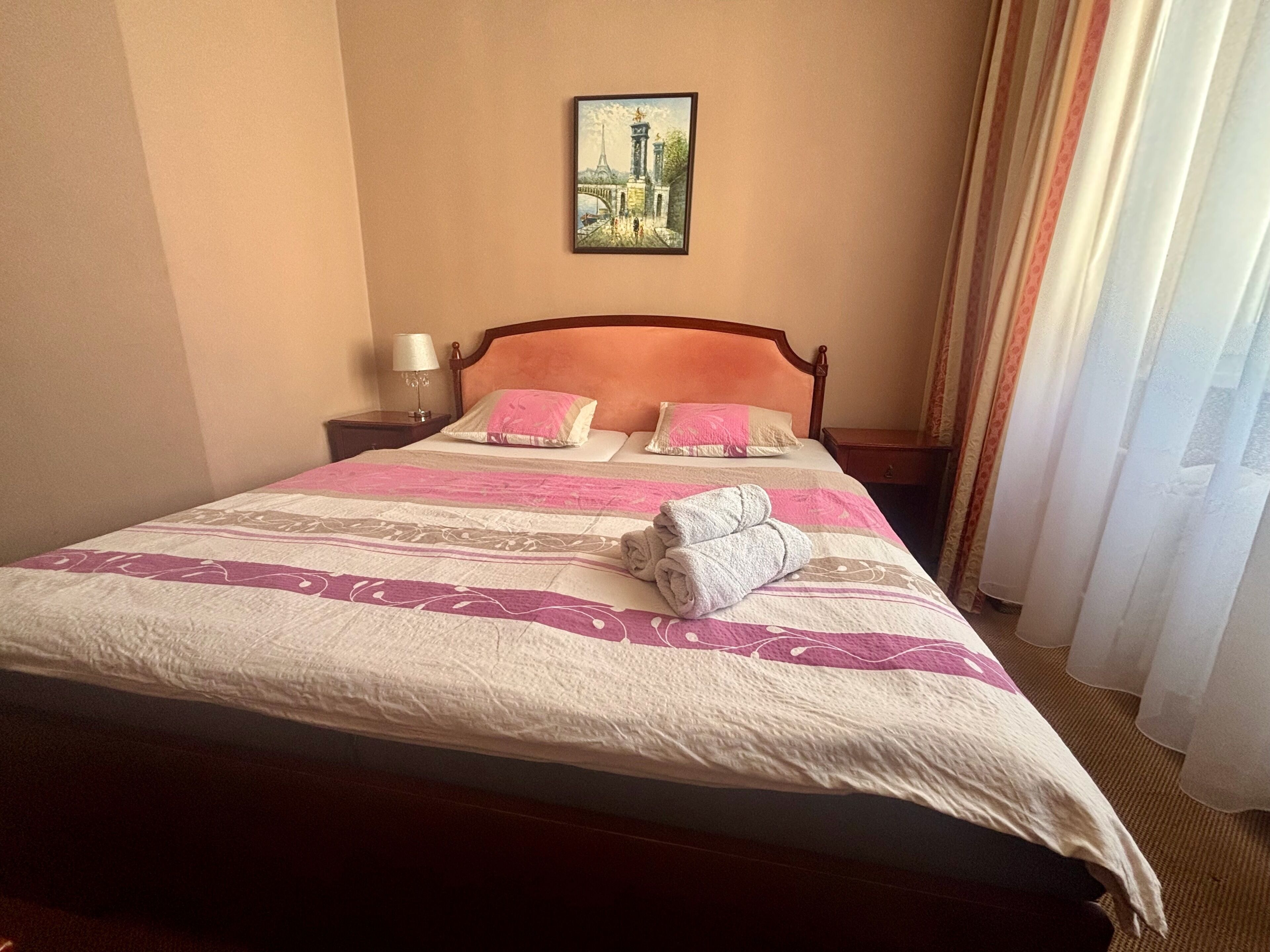 Een hotelkamer met een bed, nachtkastje, lamp, schilderij en gordijnen.