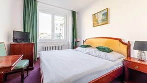 Ein Hotelzimmer mit Bett, Schreibtisch, Stuhl, Fernseher, einem Fenster mit Vorhängen, Heizkörper, einem Bild und einer Lampe.