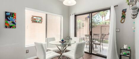 A dining area with a glass table and white chairs, a sliding glass door to an outdoor patio, and artwork on the walls.