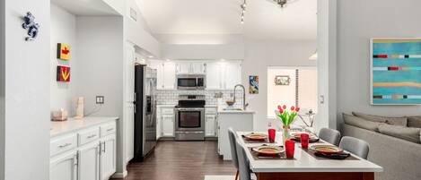 A modern kitchen with white cabinets, a dining area with a table set for four, and a living area with a sofa.