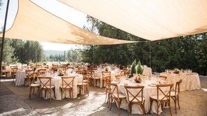 Un espacio para eventos al aire libre con mesas redondas cubiertas con manteles blancos, sillas de madera y un amplio toldo beige que proporciona sombra.