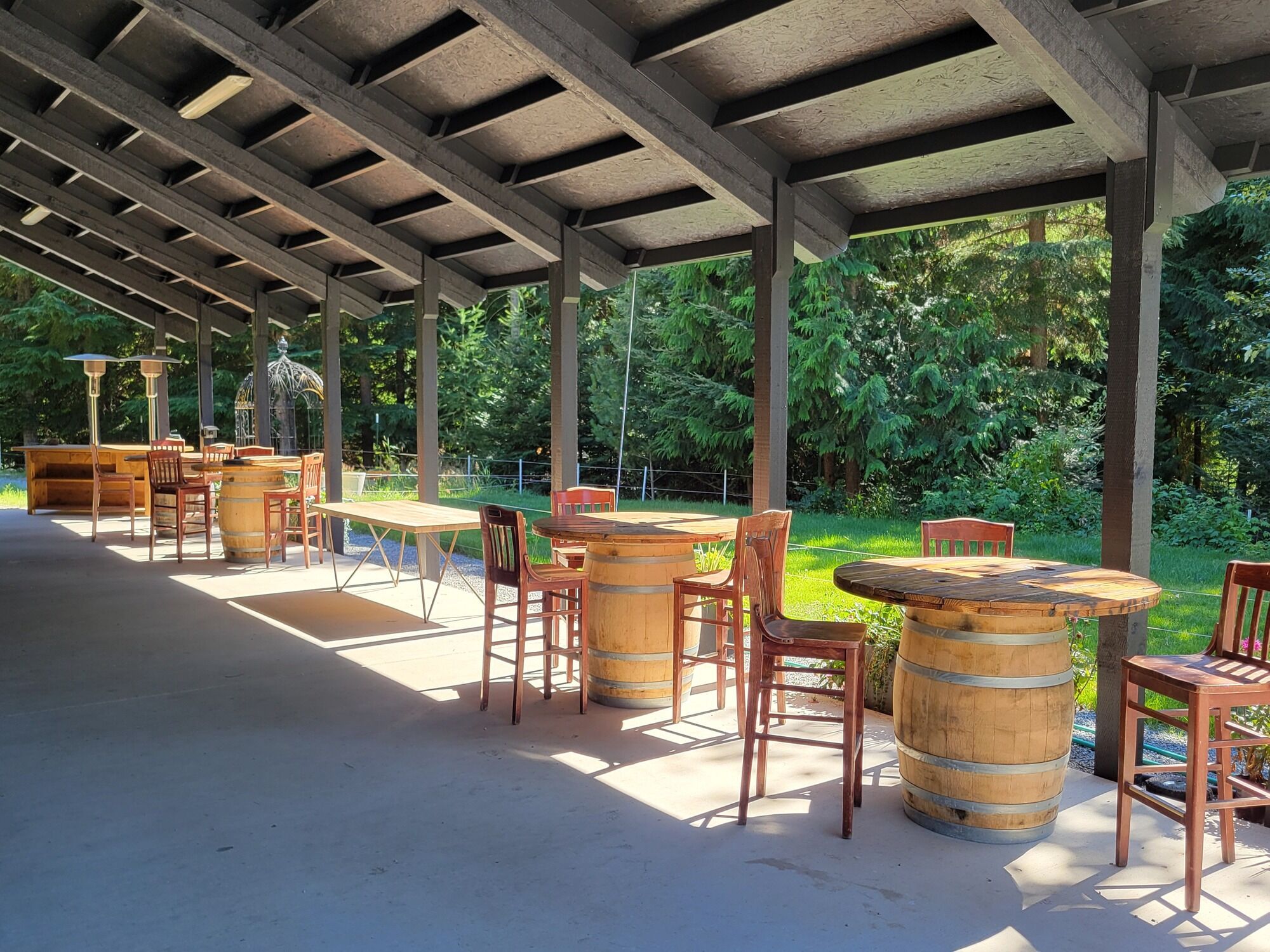 An outdoor patio with wooden tables and chairs, surrounded by greenery.