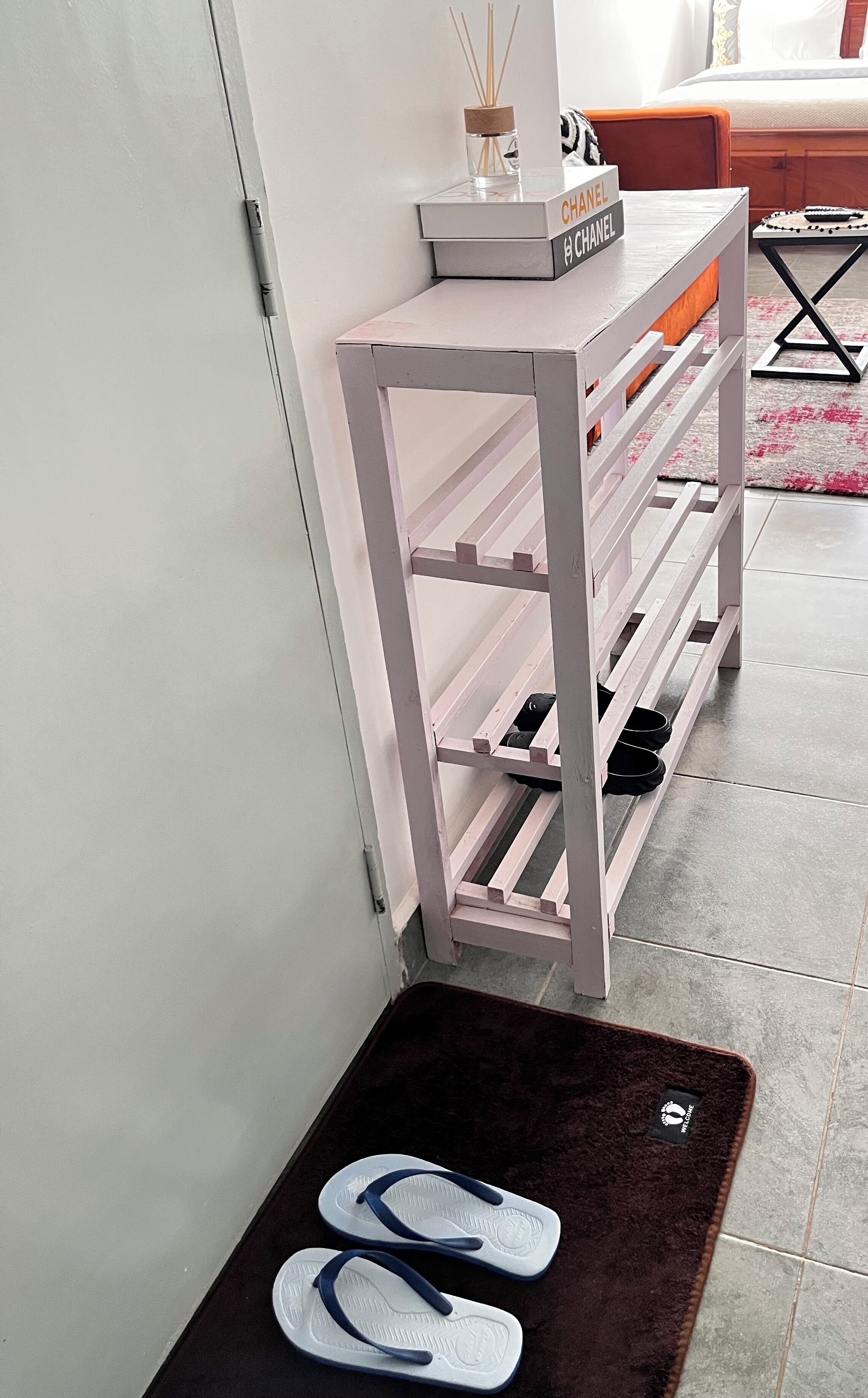 A white storage unit with shelves, a pair of flip-flops on a dark mat, and a small table with a candle and a book.