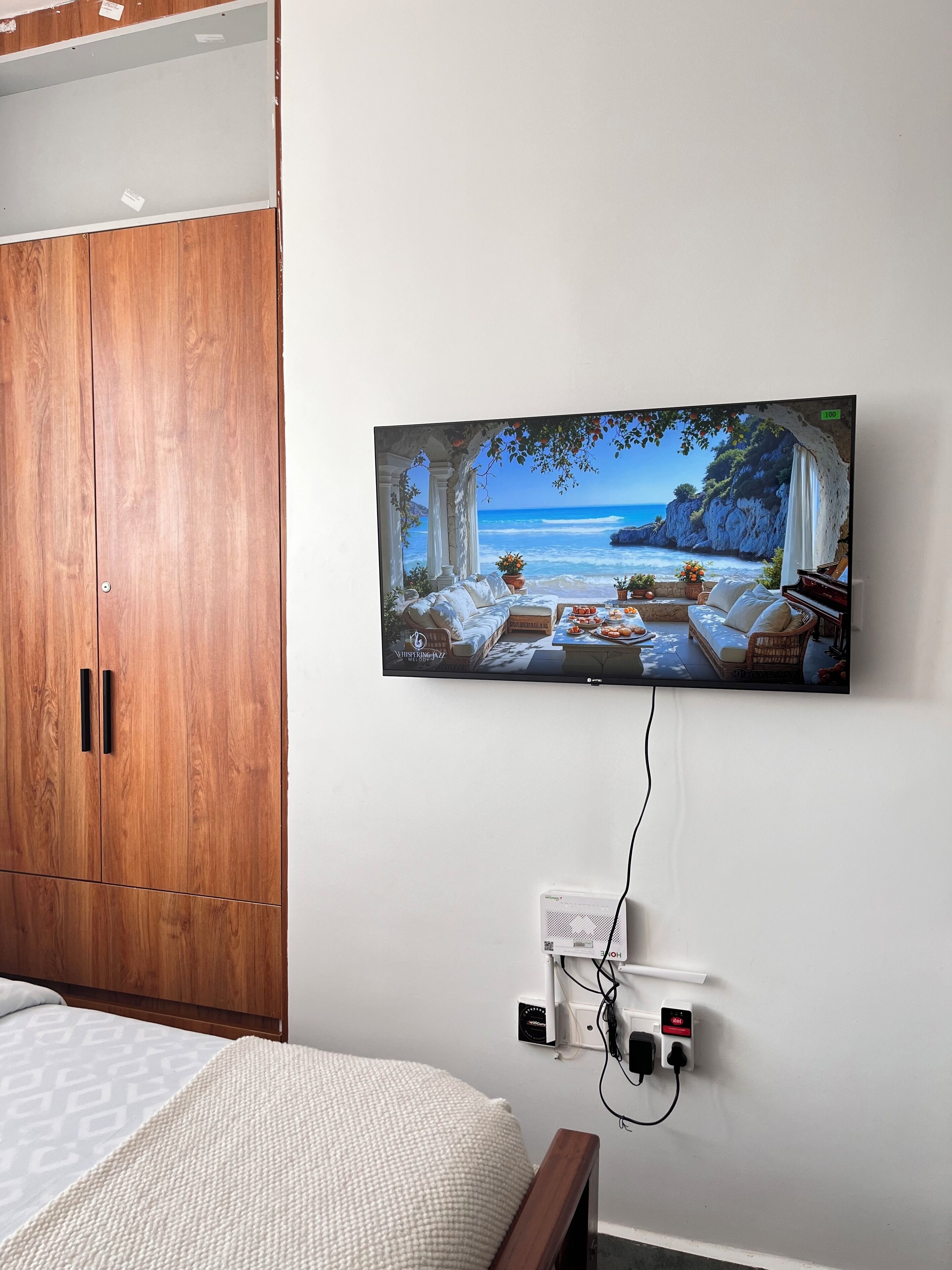 Un dormitorio con un armario de madera, una cama con manta blanca texturizada y un televisor de pared mostrando una escena de playa.