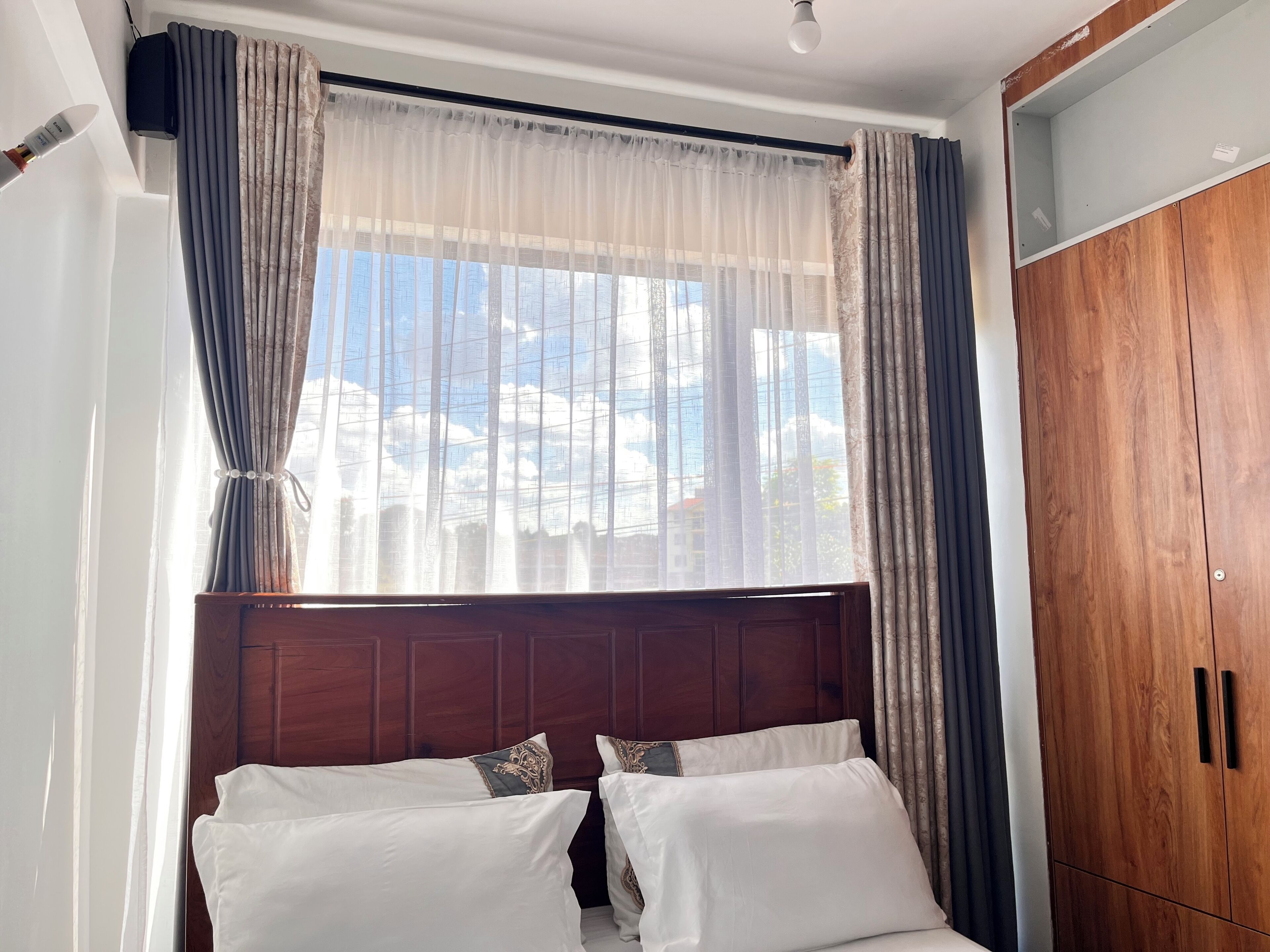Quarto com cama, cortinas, guarda-roupa de madeira e vista para edifícios pela janela.