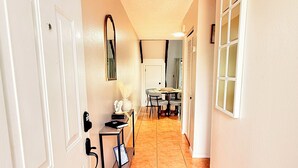 Un couloir avec une porte blanche, une petite table sur laquelle est posé un vase, et un coin repas à l’arrière-plan.