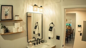 Une salle de bain moderne avec un grand miroir, un lavabo et des toilettes. On y trouve des étagères avec des articles de toilette et une plante en pot.