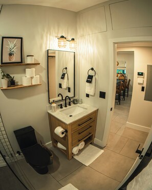 Un baño moderno con un espejo grande, un lavamanos y un inodoro. Hay repisas con artículos de aseo personal y una planta en maceta.