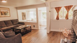 A living room with a brown sofa, a coffee table, and a fireplace. There are two windows with curtains, and a door leading outside. - Bay View Casita in Olympia (Olympia)