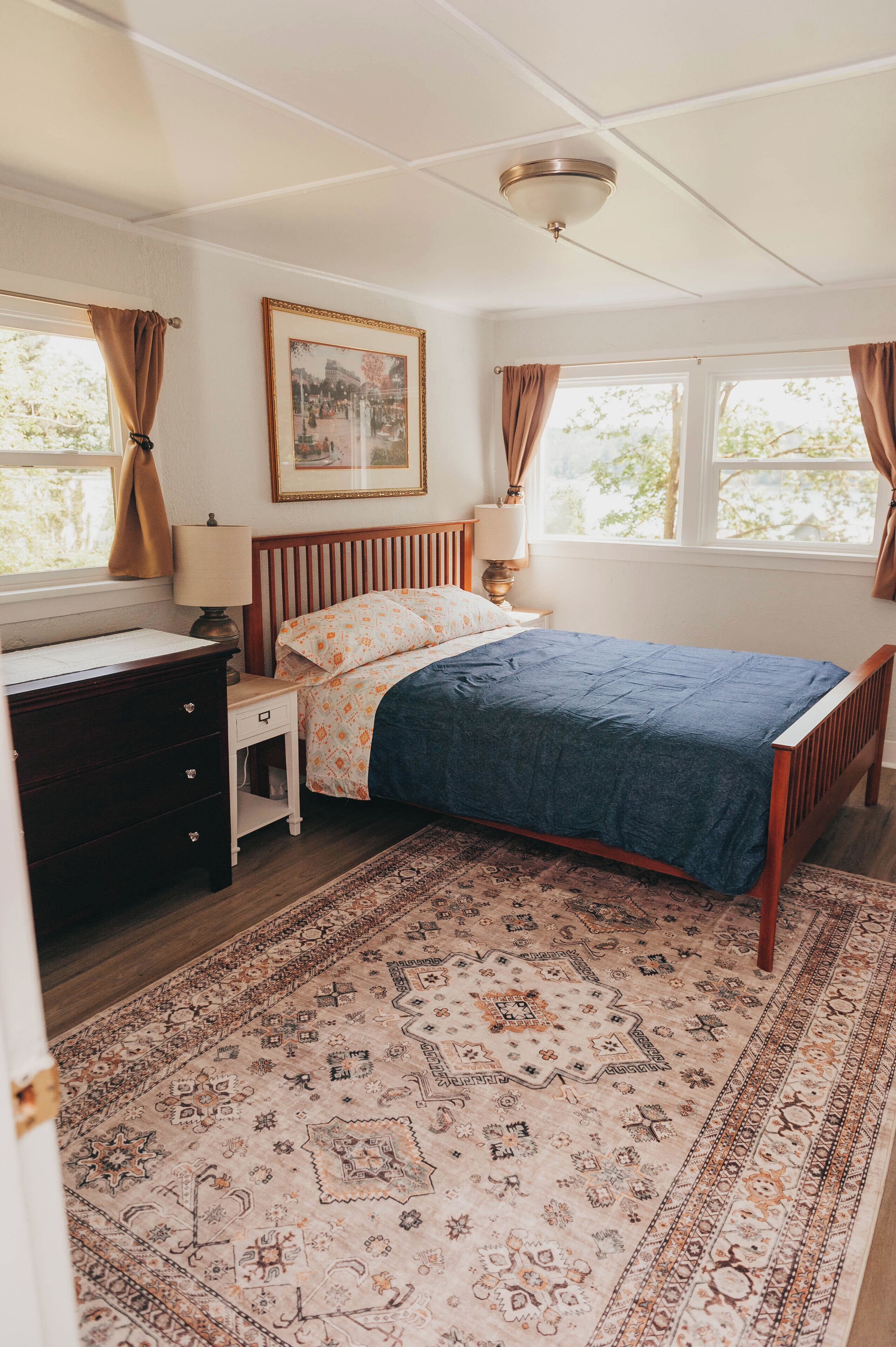 A bedroom with a wooden bed, a nightstand with a lamp, a dresser, a patterned rug, and a ceiling fan.
