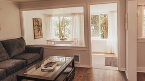A living room with a sofa, a coffee table, a window with curtains, and a door leading to an outdoor area. - Bay View Casita in Olympia (Olympia)
