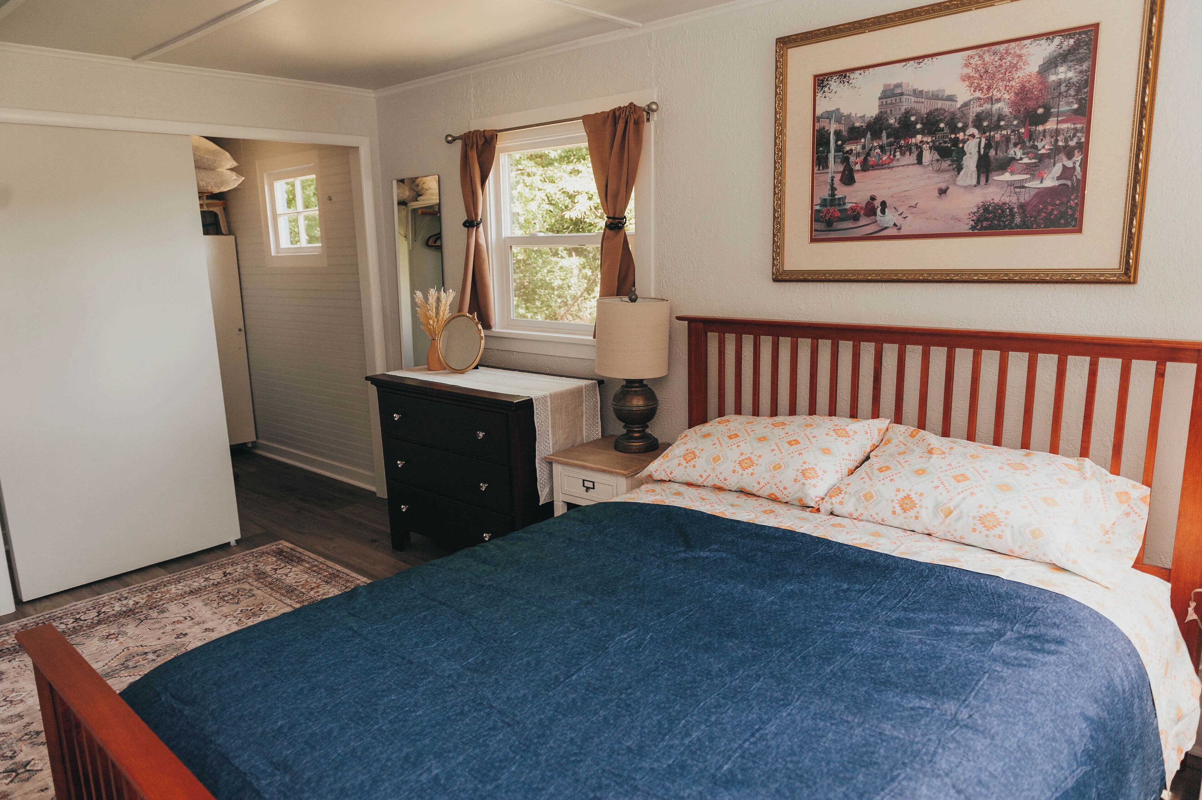 A bedroom with a bed, a nightstand, a dresser, a window with curtains, and a framed picture on the wall.