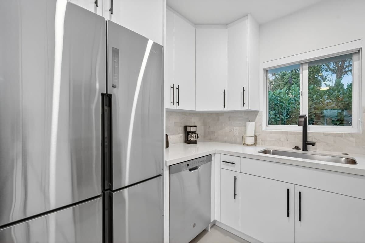 A modern kitchen with white cabinets, a stainless steel refrigerator, a dishwasher, and a window providing a view of greenery outside.
