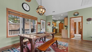 A spacious dining area with a wooden table and benches, a large window with blinds, a chandelier, and a kitchen in the background. - Fairway & Mountain Views, Relaxing 2BR Condo Retreat (Whittier)