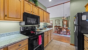 A kitchen with wooden cabinets, a black microwave, and a stove. There is a dining area with a table and chairs. - Fairway & Mountain Views, Relaxing 2BR Condo Retreat (Whittier)