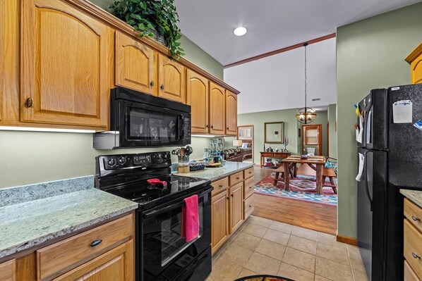 A kitchen with wooden cabinets, a black microwave, and a stove. There is a dining area with a table and chairs. - Fairway & Mountain Views, Relaxing 2BR Condo Retreat (Whittier)