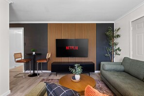Ein modernes Wohnzimmer mit Flachbildfernseher, einer Couch, einem Couchtisch und einer kleinen Essecke mit Stühlen.