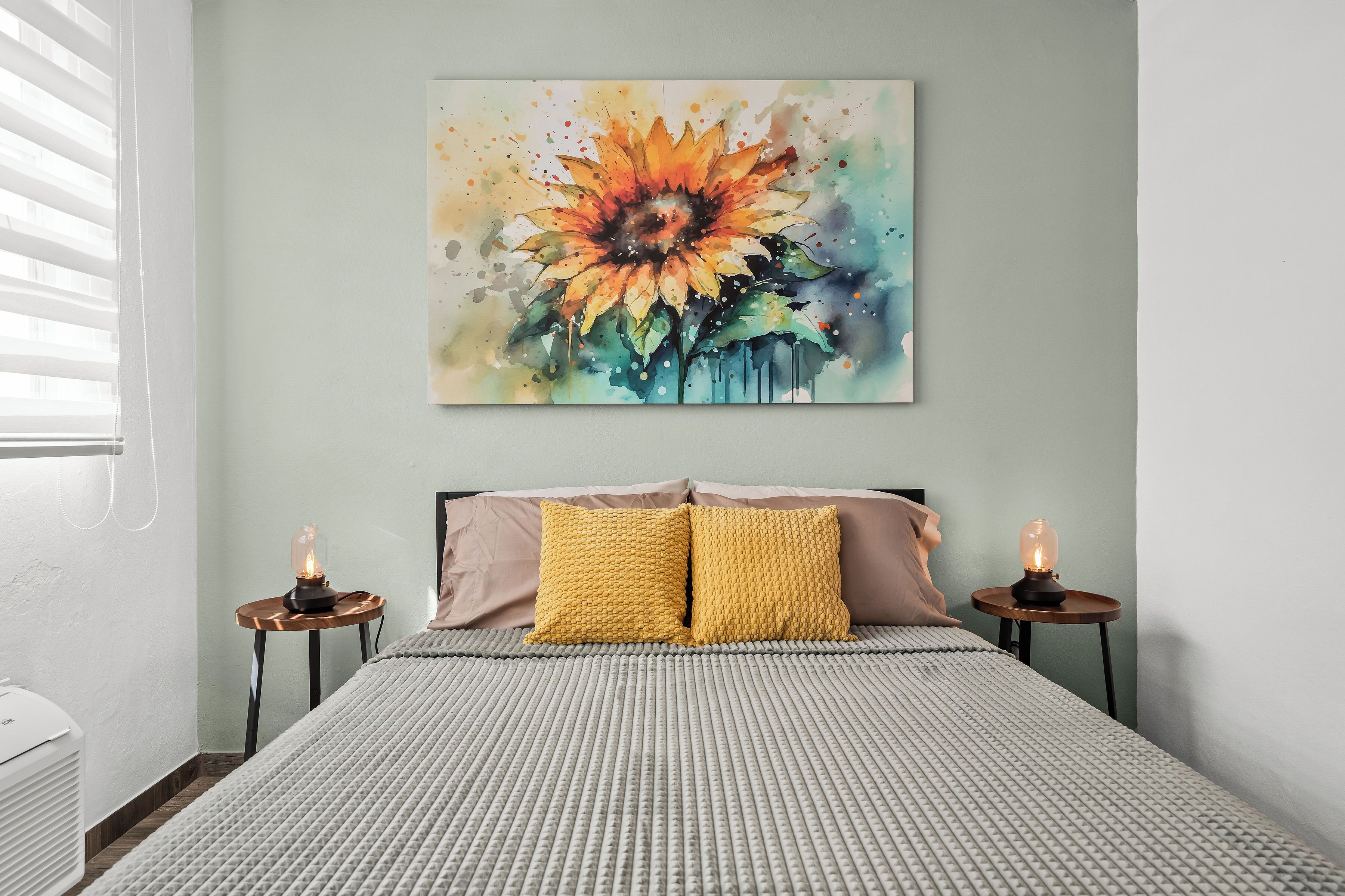 Une chambre à coucher avec un lit, deux tables de chevet avec des lampes, un tableau représentant un tournesol accroché au mur, et une fenêtre avec des stores.