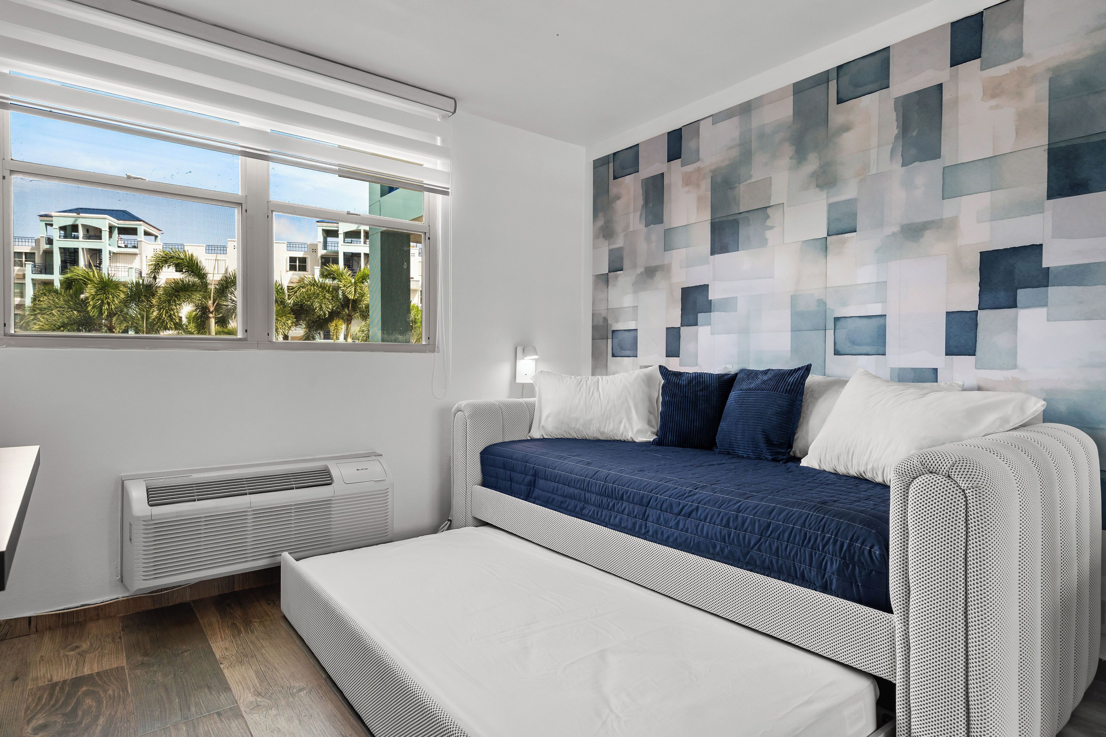 A modern bedroom with a bed, a bench, a wall-mounted air conditioner, and a window with a view of buildings and palm trees.