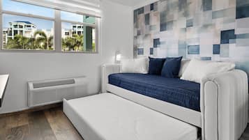 A modern bedroom with a bed, a bench, a wall-mounted air conditioner, and a window with a view of buildings and palm trees.