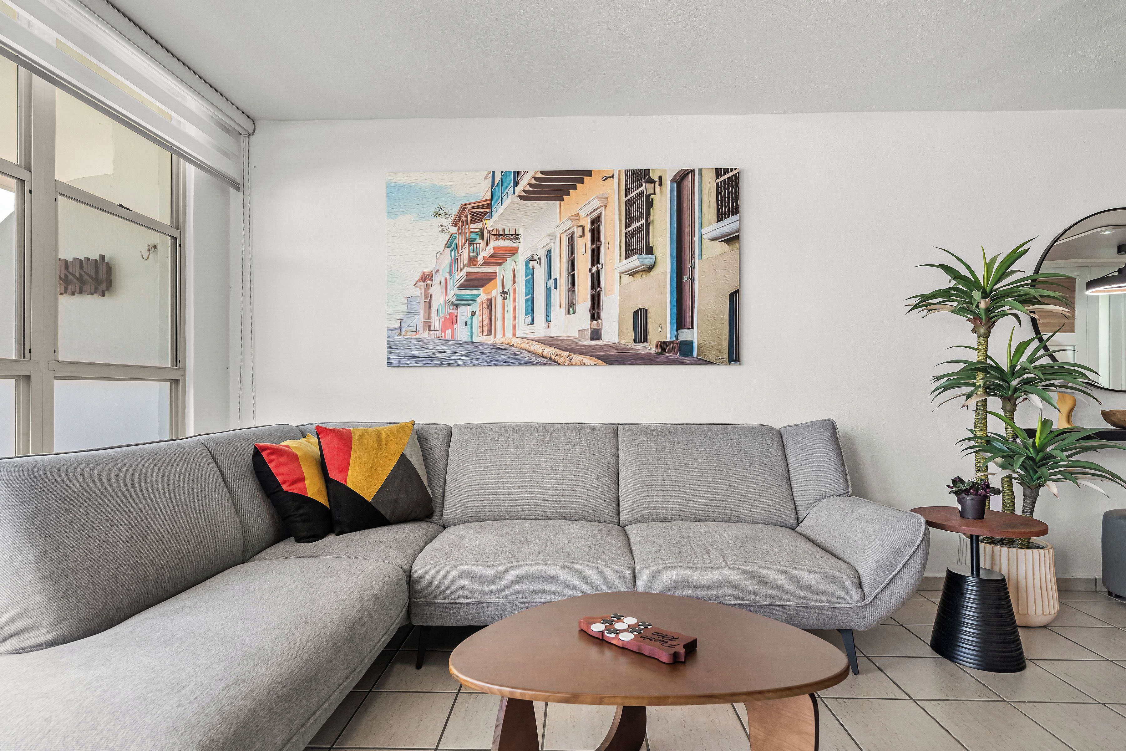 Un salon moderne avec un canapé d’angle gris, une table basse, un tableau représentant une scène de rue et deux plantes en pot.